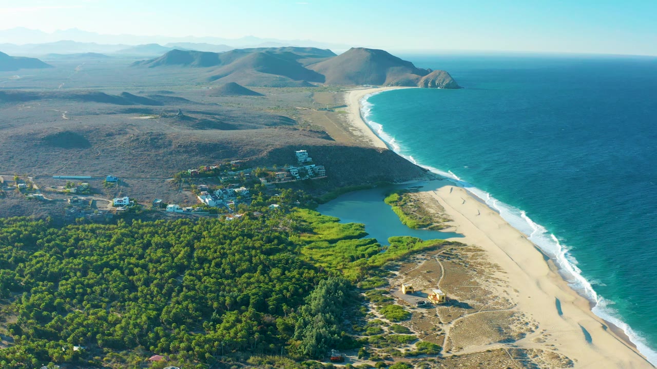 Drone-height view over Todos Santos coastline with turquoise ocean on the right, a pale beach curving past a small lagoon, dense greenery below, scattered buildings inland, and no visible people.