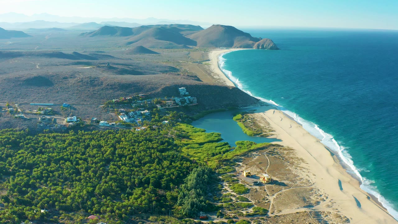 Wide aerial frame in Todos Santos showing blue ocean and white surf beside a long sandy beach, green trees and a small developed area inland, dry mountains in the distance, and no visible people.