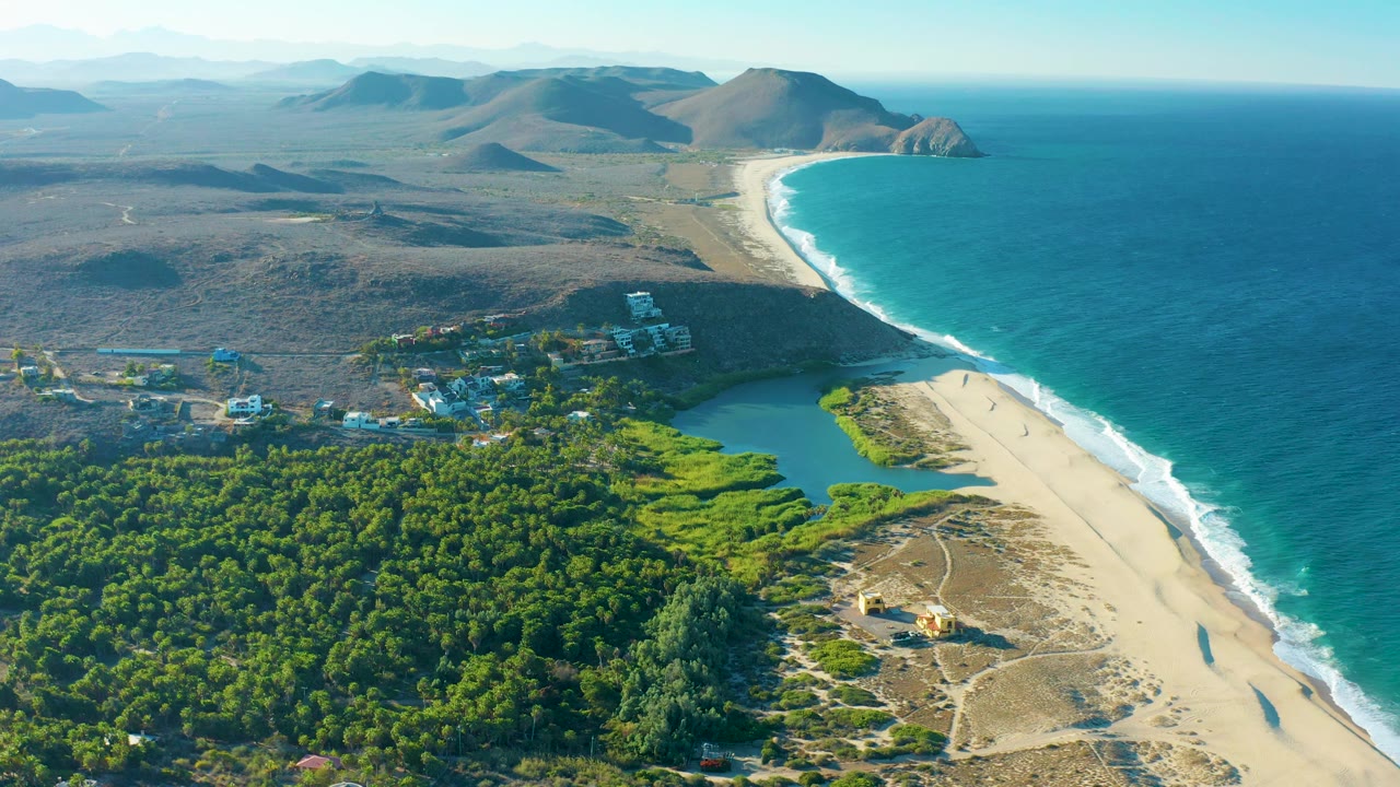 High coastal aerial with forest foreground, lagoon, beach, waves, and hills