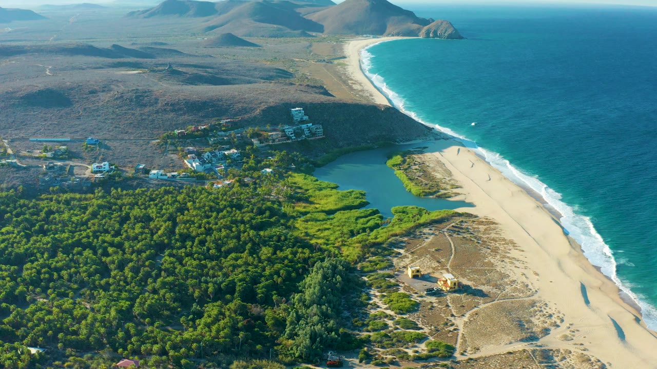 Aerial view of Todos Santos coastline with turquoise ocean, white surf, a sandy beach, green forest, lagoon water, scattered village buildings, dry hills, and no visible people.