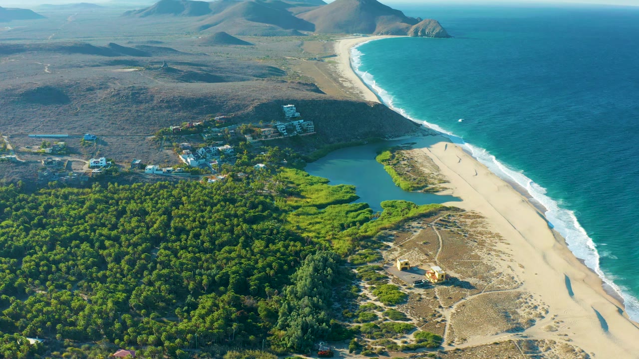 High aerial frame over Todos Santos with the surf line running along the right edge, lagoon and village buildings near center, forest in the lower left, hills in back, and no visible people.