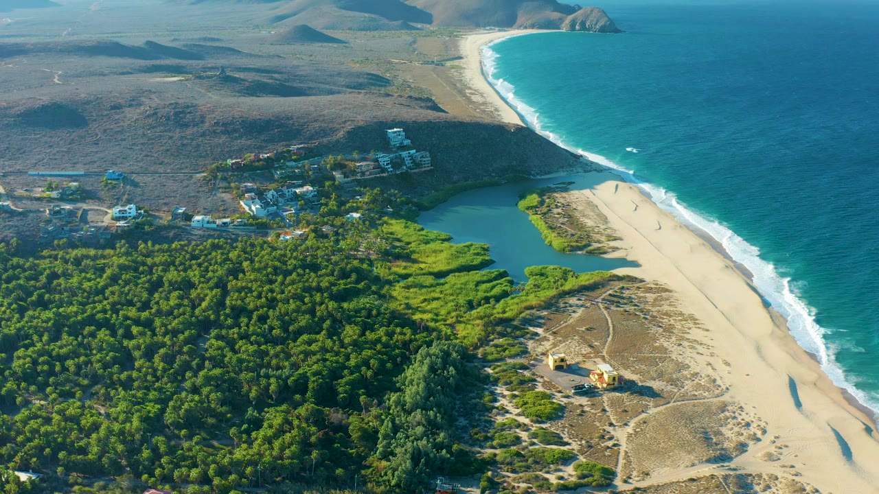 Aerial beach landscape with shadowed hills, forest, lagoon, and ocean surf.