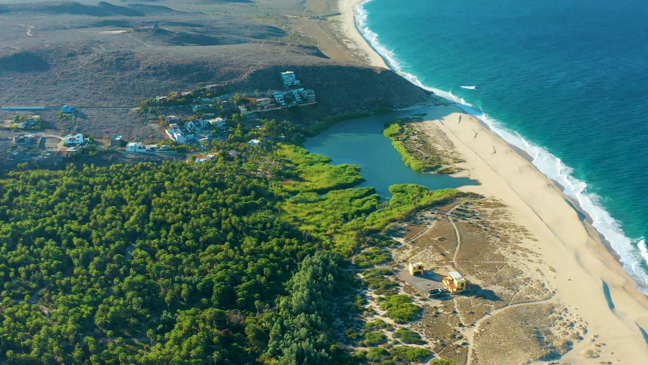 Aerial view over Todos Santos, Baja California Sur, with dense green forest at lower left, a lagoon near the beach, scattered buildings, small beach walkers, and blue ocean surf at right.