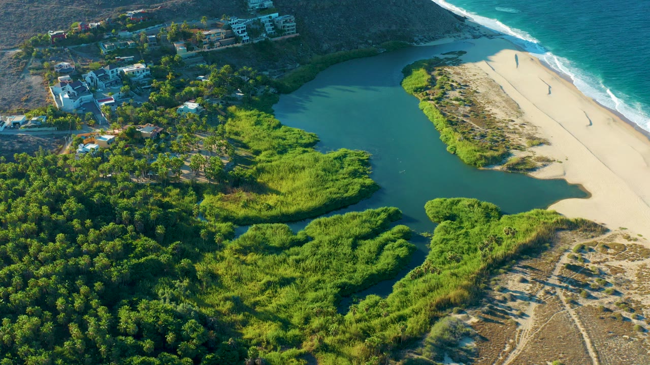 Aerial frame from Todos Santos, Baja California Sur showing green lagoon water beside a pale sand beach, ocean surf at the top edge, dense vegetation, hillside homes, and no visible people.
