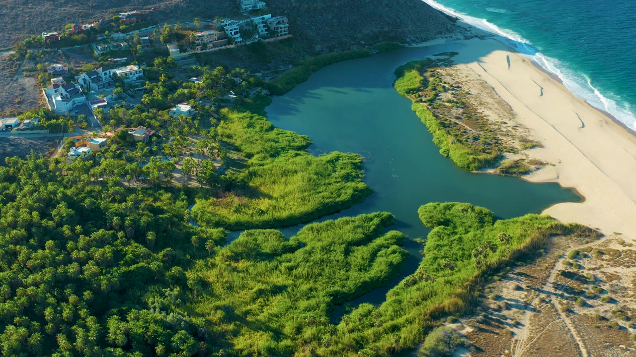 Aerial frame from Todos Santos, Baja California Sur showing a wider sweep of lagoon water, sandbar, beach, surf, thick green vegetation, scattered homes near the hillside, and no visible people.