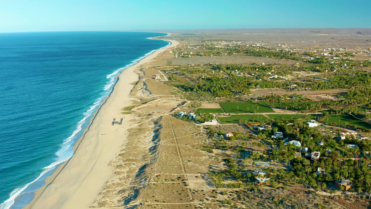 Opening aerial frame over Todos Santos, Baja California Sur, with blue ocean at left, a long sandy beach, dunes, green fields, scattered buildings, and no visible people.