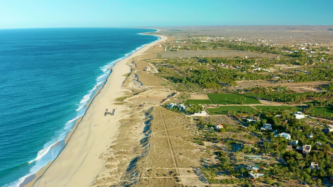 Aerial view over Todos Santos, Baja California Sur, with blue ocean on the left, a long sandy beach and dunes through the center, inland green fields and scattered buildings to the right, and no visible people.