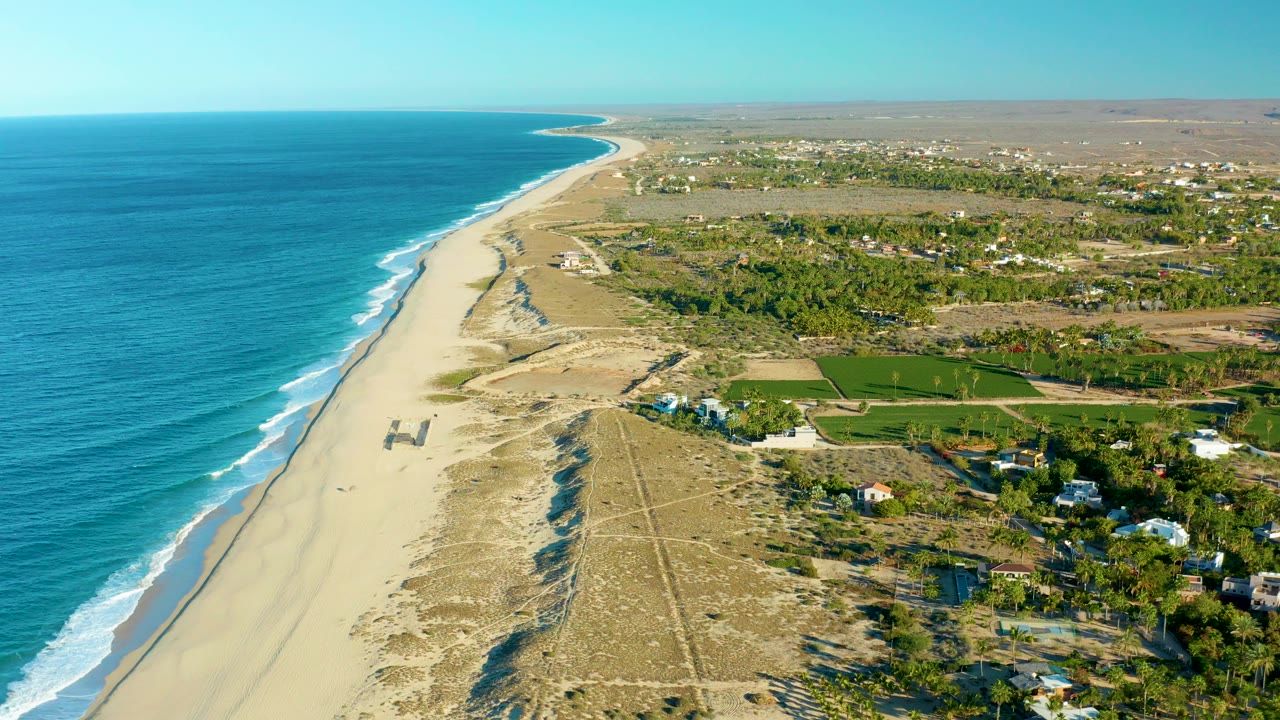 Aerial view of Todos Santos, Baja California Sur with blue ocean at left, a sandy beach and dunes below, inland green fields and scattered houses to the right, and no visible people.