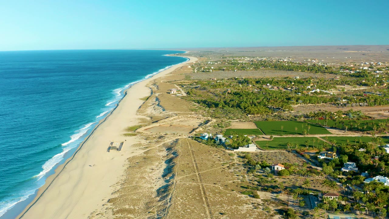 Opening aerial view over Todos Santos, Baja California Sur, with blue ocean on the left, a wide sandy beach, dune tracks, inland green fields, scattered buildings, and no visible people.