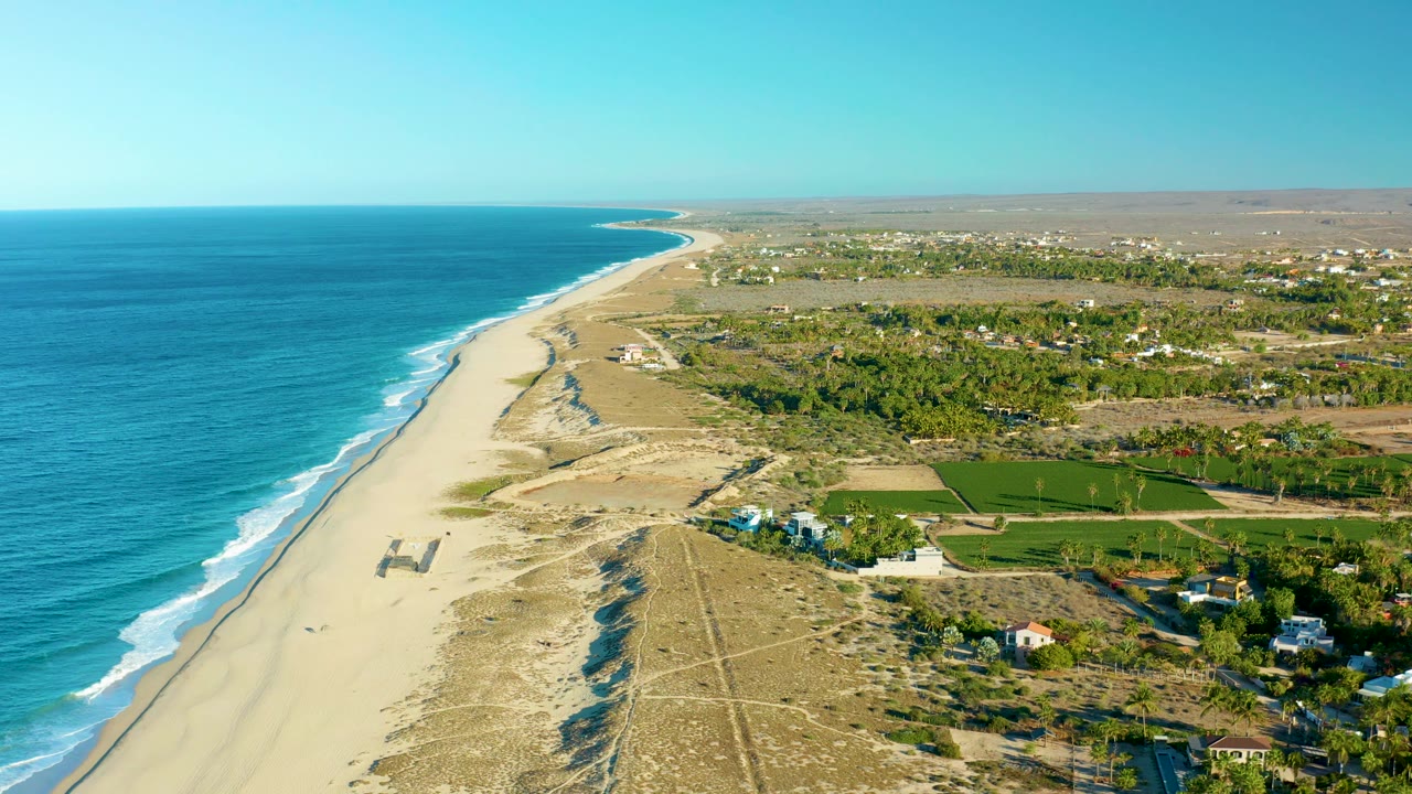 Aerial view of sandy shoreline with ocean at left and green fields near scattered houses.