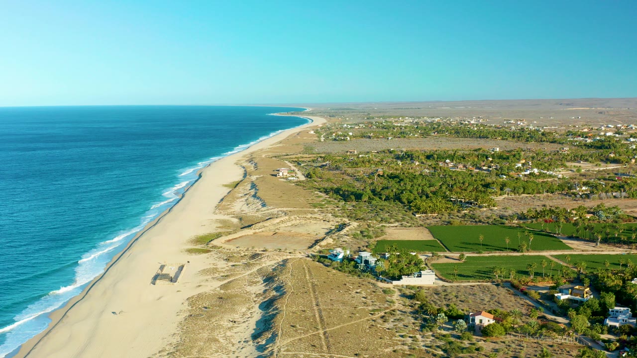 High aerial view over Todos Santos, Baja California Sur, showing blue ocean on the left, a long sandy beach and dunes through the center, inland green fields and scattered buildings, and no visible people.