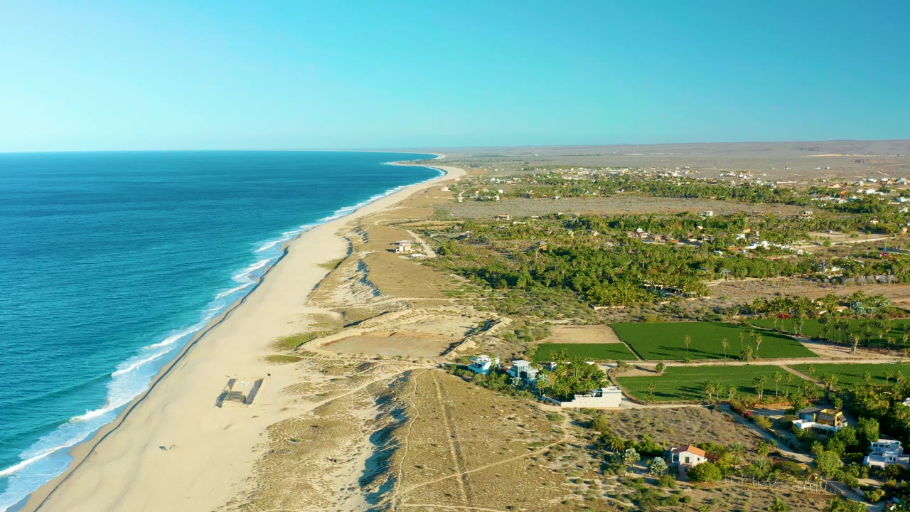 Aerial frame of Todos Santos coastline with the surf line slightly farther along the beach, dry dunes in the foreground, green rectangular fields inland, scattered structures, and no visible people.