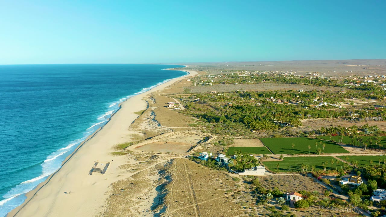 Wide drone view over Todos Santos with turquoise-blue water, thin white wave lines, a broad pale beach, tan dunes, green fields to the right, scattered inland buildings, and no visible people.