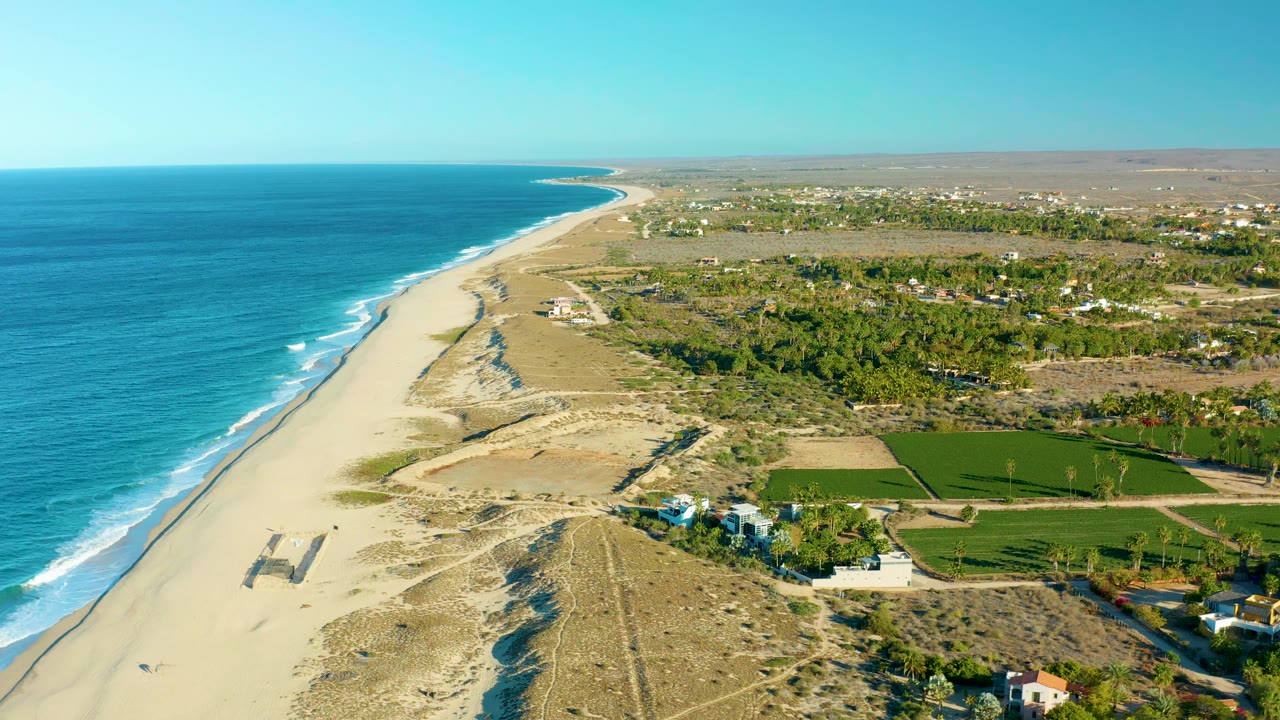 High aerial view in Todos Santos showing the Pacific shoreline, pale sand, low dunes, green fields, inland buildings, clear sky, and no visible people.