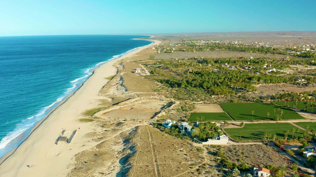 Aerial beach scene with turquoise water, surf, dunes, fields, and coastal buildings.