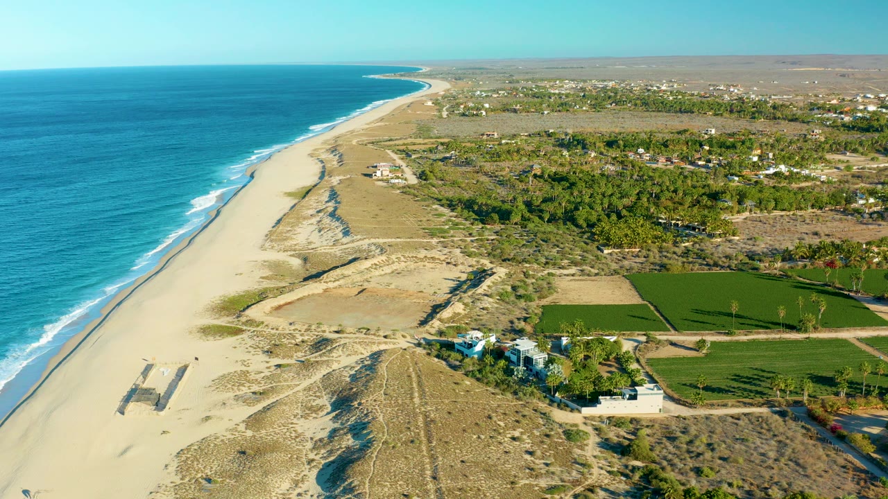 Opening aerial frame over Todos Santos, Baja California Sur, with blue ocean at left, pale sand beach, dunes, green fields, and small white buildings inland, with no visible people.