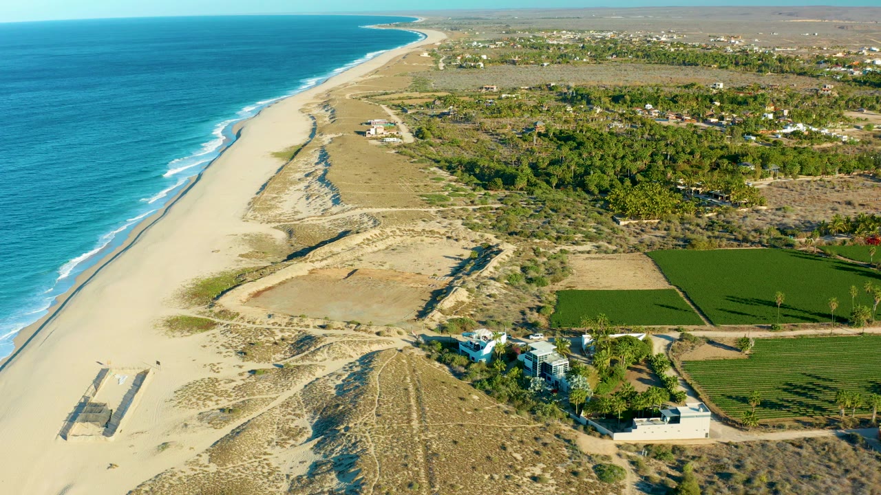 Opening aerial frame over Todos Santos, Baja California Sur, with blue ocean along the left edge, pale sand and dunes through the center, green fields inland, a coastal house construction site near the lower right, and no visible people.