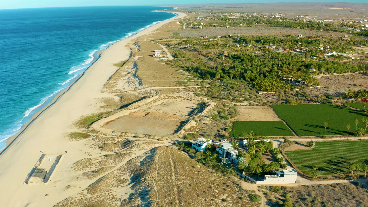 At 0:01, the aerial view in Todos Santos, Baja California Sur keeps the shoreline running diagonally, with a rectangular structure on the sand at lower left, construction near the lower right, inland fields, trees, and no visible people.