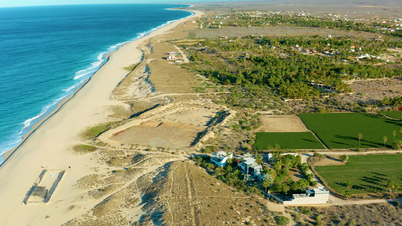 At 0:02, the Todos Santos coastline remains in a high oblique view, with turquoise water and surf on the left, broad sand and dunes in the middle, a cluster of coastal buildings near the lower right, fields inland, and no visible people.