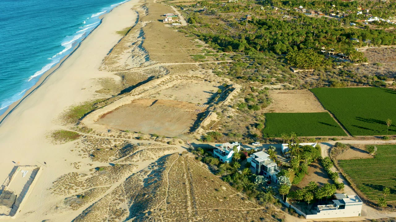 Aerial view in Todos Santos with blue water and breaking waves at left, open sand and dunes across the middle, farm fields on the right, small buildings near the lower edge, and no visible people.