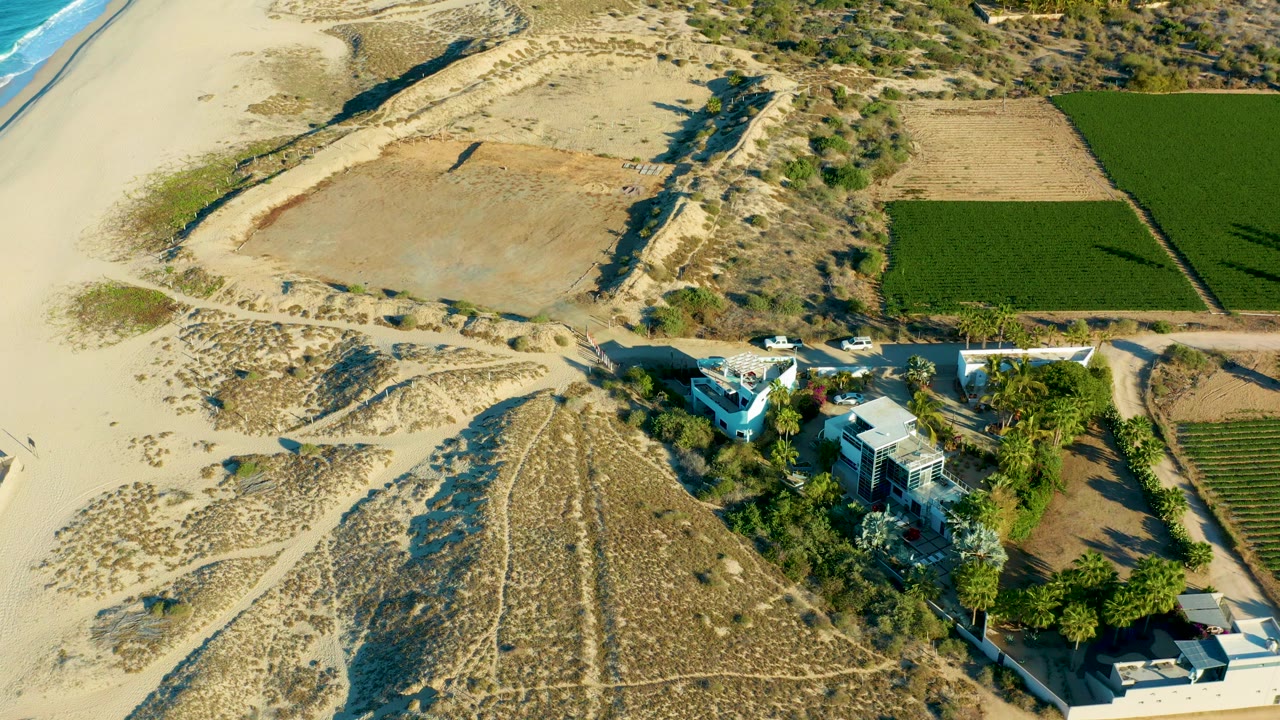 High overhead view in Todos Santos with the white car farther along the road beside sandy dunes, a bright green rectangular field, dry farmland, houses, and no visible people.