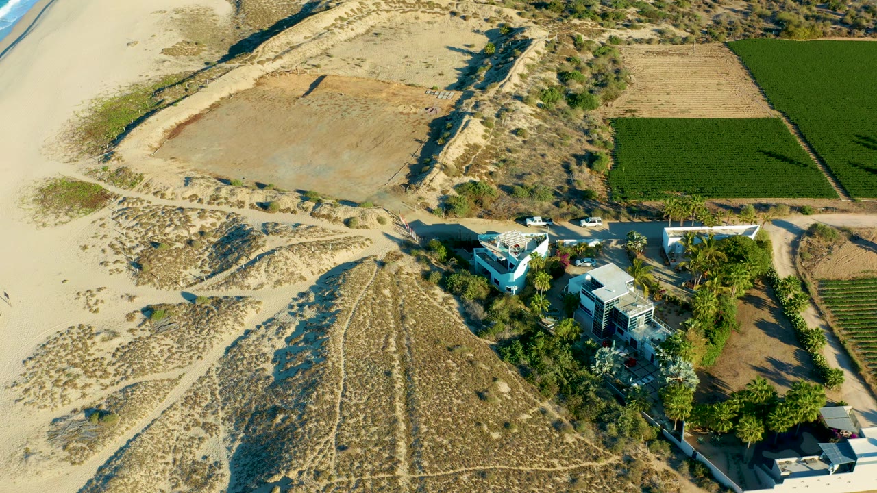 Aerial frame over Todos Santos where the ocean edge enters the upper left, with pale sand, a looping dune track, houses, trees, green farmland, and no visible people.