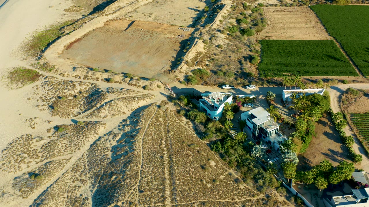 Aerial view of a white car moving near houses between dunes and a green field.