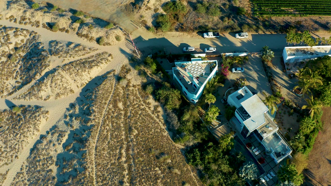 Overhead frame in Todos Santos, Baja California Sur showing sandy dunes, a roadside with parked vehicles, flat-roofed buildings, palms, and no visible people.
