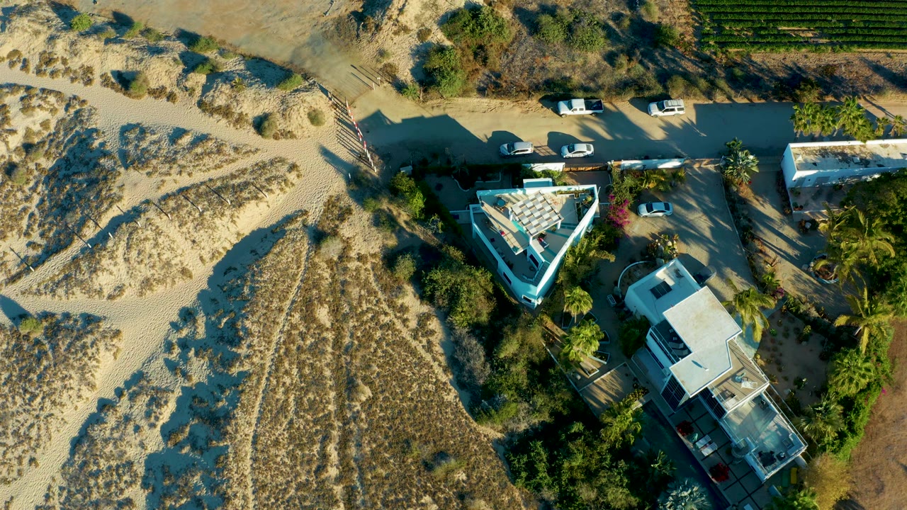 Overhead Todos Santos frame showing dunes below a roadside with parked vehicles, residential structures among palms, a green agricultural field at the upper right, and no visible people.