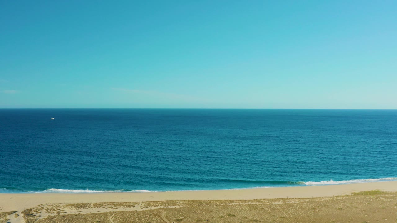 Aerial frame over Todos Santos, Baja California Sur showing blue ocean, a pale sandy beach, low scrub at the bottom edge, a small distant boat on the water, and no visible people.