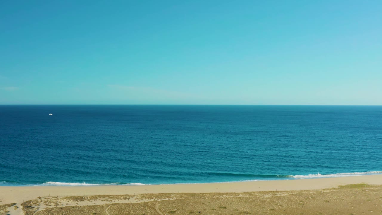 Aerial view in Todos Santos with turquoise water across the middle of frame, pale sand along the lower third, scrubby dunes below, a tiny distant boat near the horizon area, and no visible people.