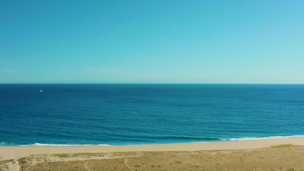 Todos Santos aerial frame with a broad blue sea, thin white surf at the shoreline, tan beach, sparse vegetation, a small white boat far offshore, and no visible people.