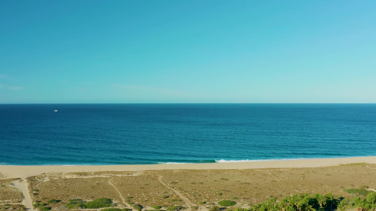 Aerial view of blue ocean, sandy beach, low dunes, and a tiny boat offshore.