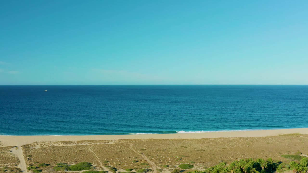 Todos Santos aerial view with the horizon high in frame, pale beach and scrub-covered dunes below, and the small offshore boat shifted farther across the water, with no visible people.