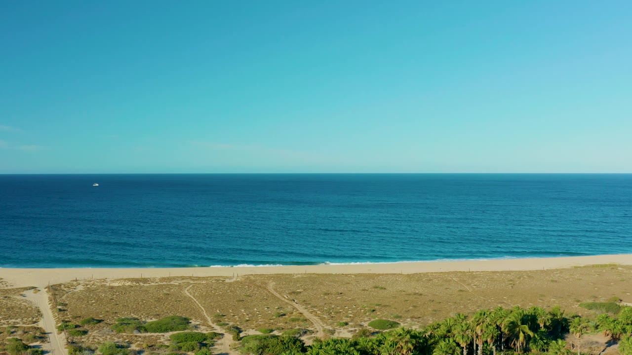Todos Santos coastline frame showing broad sky, calm ocean, pale sand, scrubby dunes, and a tiny boat left of center on the water, with no visible people on the beach.
