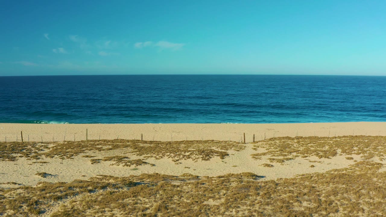 Aerial frame from Todos Santos showing calm blue ocean, a thin band of white surf, broad sand, dry dune vegetation, and no visible people.
