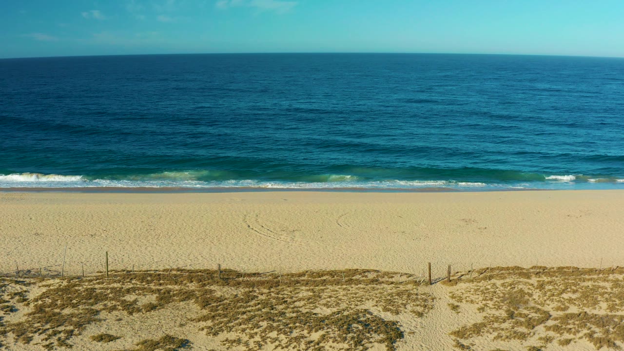High view over a Todos Santos beach where blue water approaches a pale shoreline, with low dunes and small fence posts visible below and no visible people.