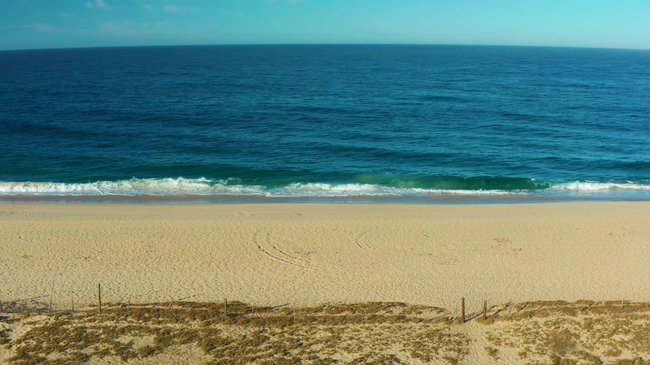 Todos Santos beach frame with the camera looking over pale sand toward blue ocean as a small wave breaks near shore, with no visible people.