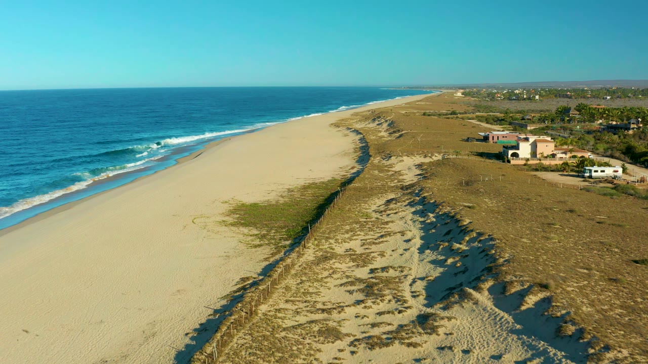 Aerial view in Todos Santos with the ocean and breaking waves along the left edge, a pale sandy beach below, dunes casting shadows, and low buildings near the road on the right, with no visible people.