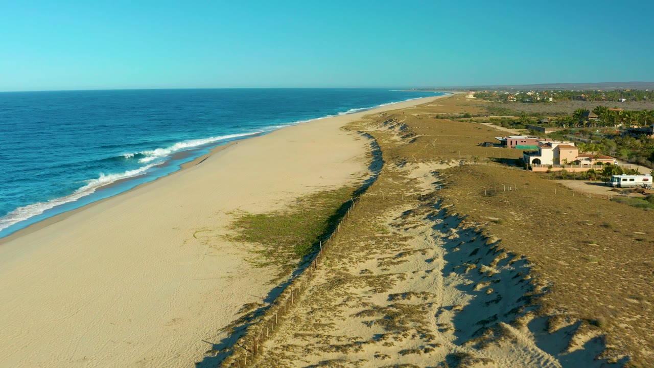 Elevated Todos Santos coastal frame showing the surf band on the left, uninterrupted sand in the middle, dune vegetation and shadow patterns to the right, and several roadside structures beyond, with no visible people.