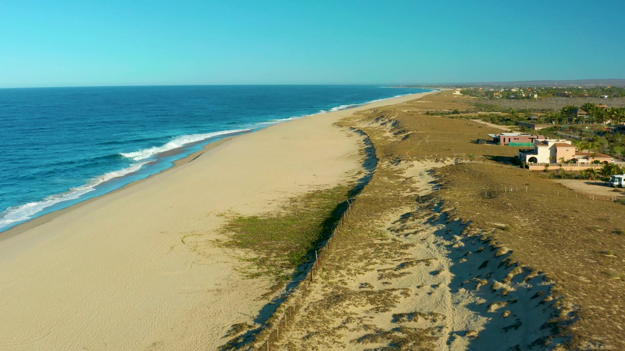Aerial beach view with ocean waves, dune grass, road, buildings, and a white van.