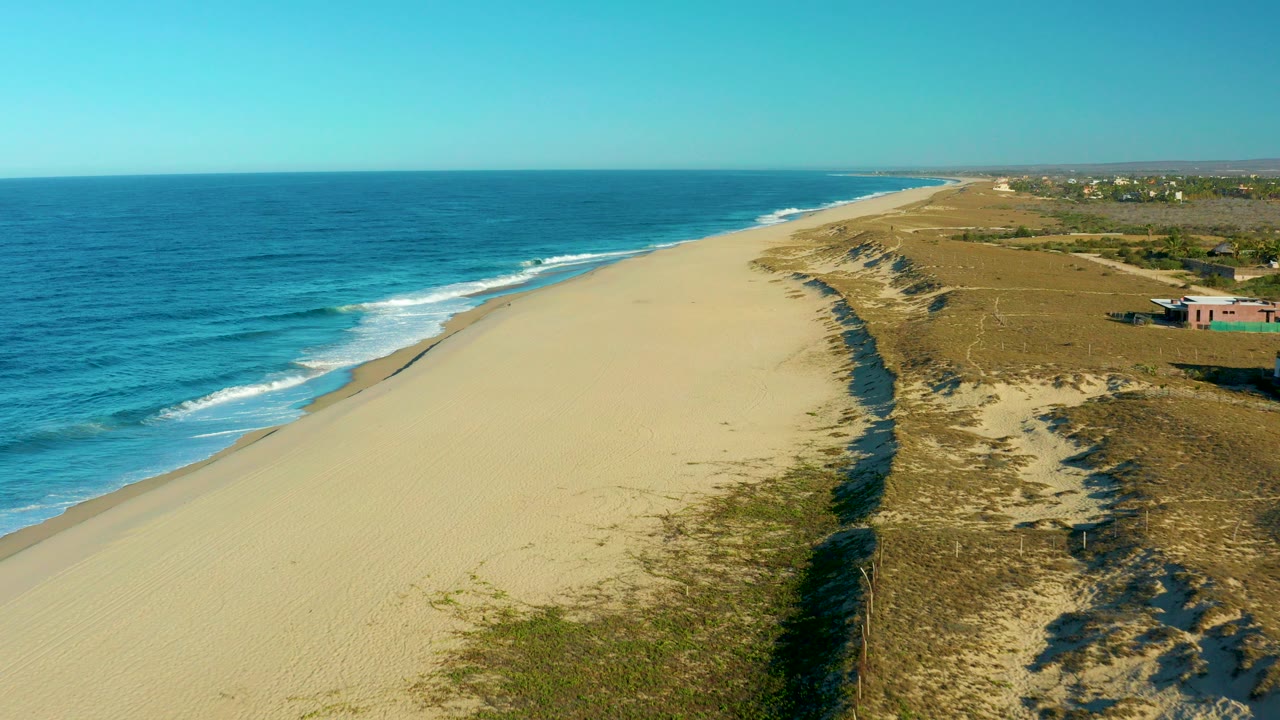 Aerial view over Todos Santos, Baja California Sur, with blue ocean on the left, a wide sandy beach through the center, low dunes on the right, and no visible people.