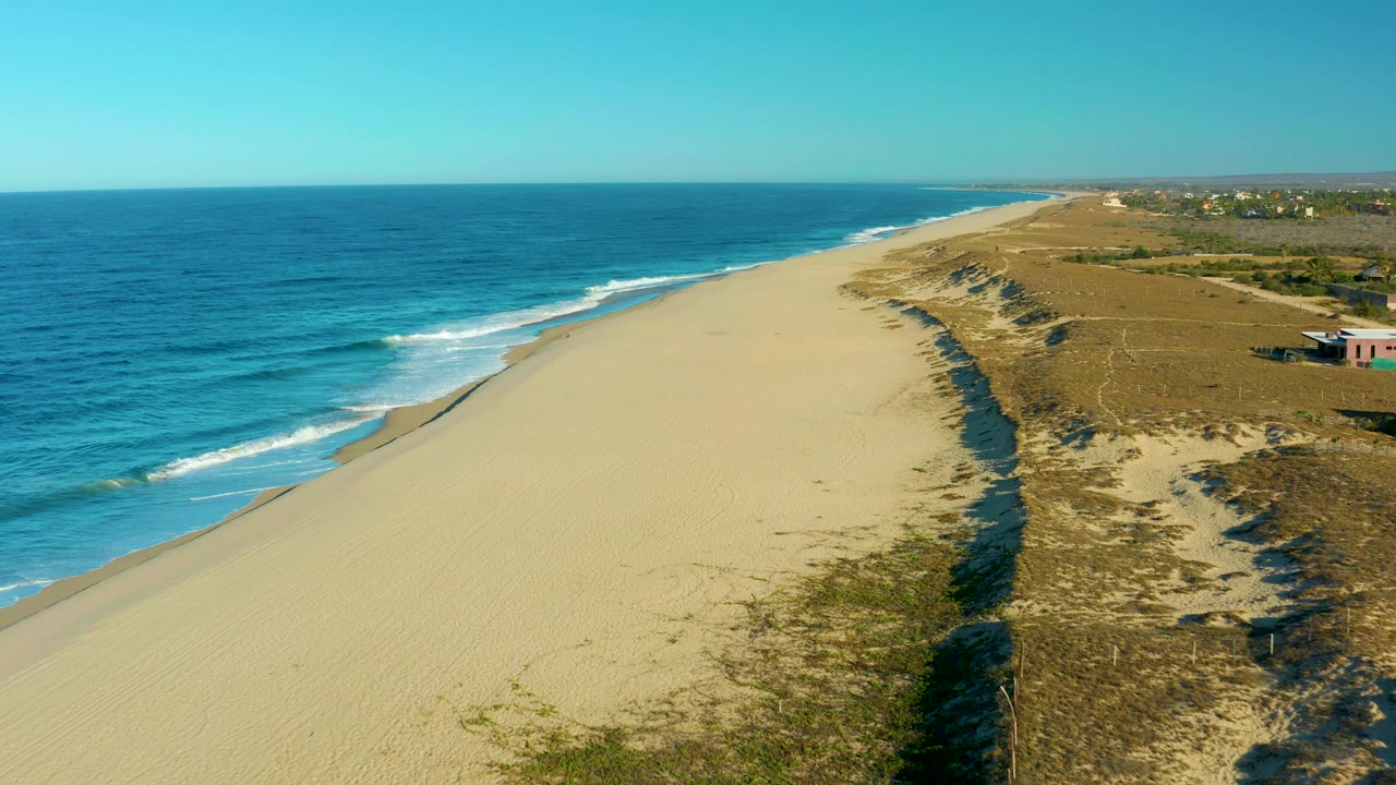 Aerial view along the Todos Santos coast with the shoreline curving into the distance, ocean occupying the left half, dunes and sparse buildings to the right, and no visible people.