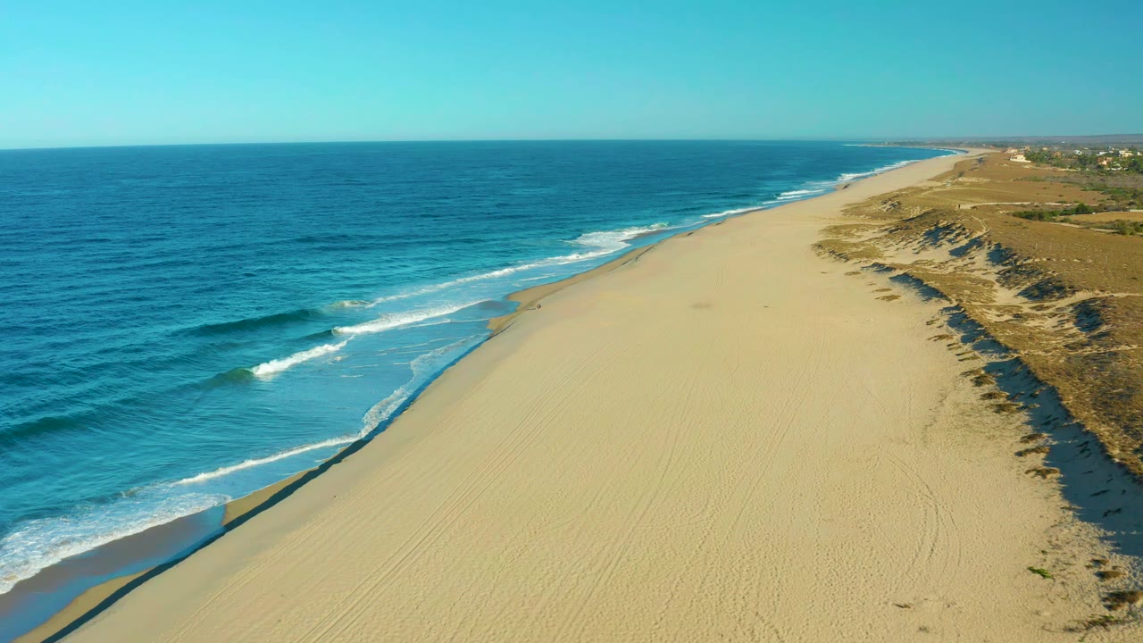 Aerial frame in Todos Santos, Mexico, showing white surf curling along the left edge of a tire-marked sandy shoreline, with dunes and sparse vegetation on the right and no visible people.