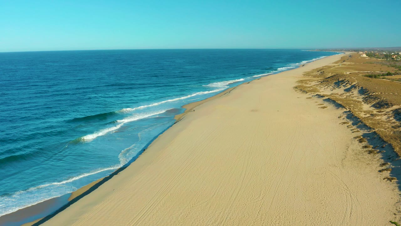 High oblique aerial over Todos Santos, Mexico, with a darker blue swell approaching pale sand, foam bands near shore, dune ridges to the right, and no visible people.