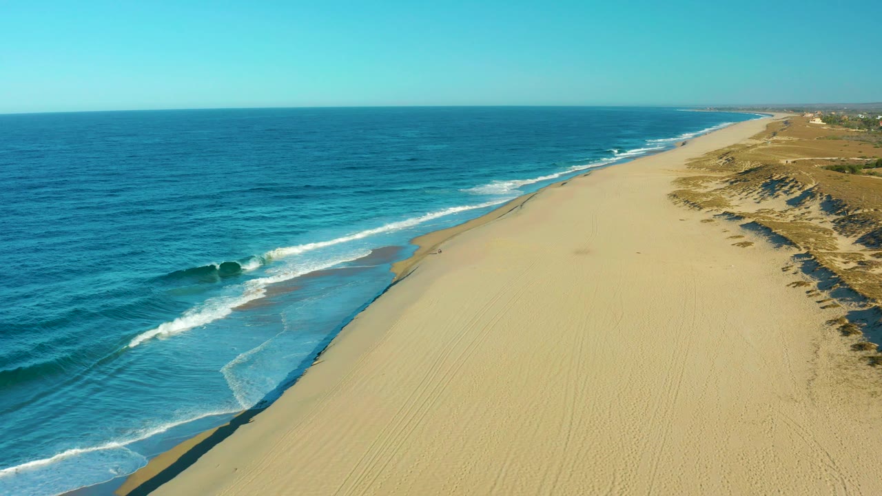 Aerial view in Todos Santos, Mexico, where white water spreads across wet sand beside a broad empty beach, with low dunes and vegetation continuing along the right side and no visible people.
