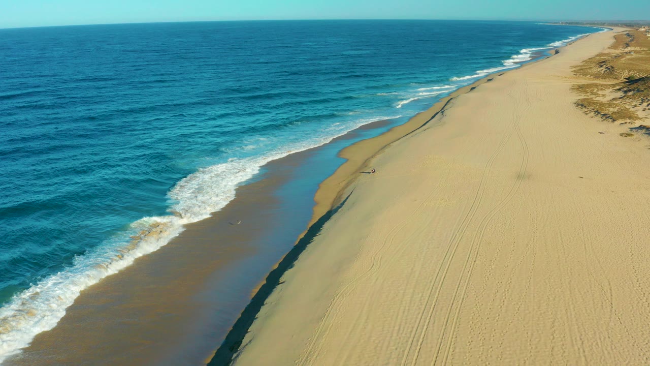 Aerial view in Todos Santos, Baja California Sur, with deep blue ocean on the left, tan sand on the right, and a small person walking near the wet shoreline.