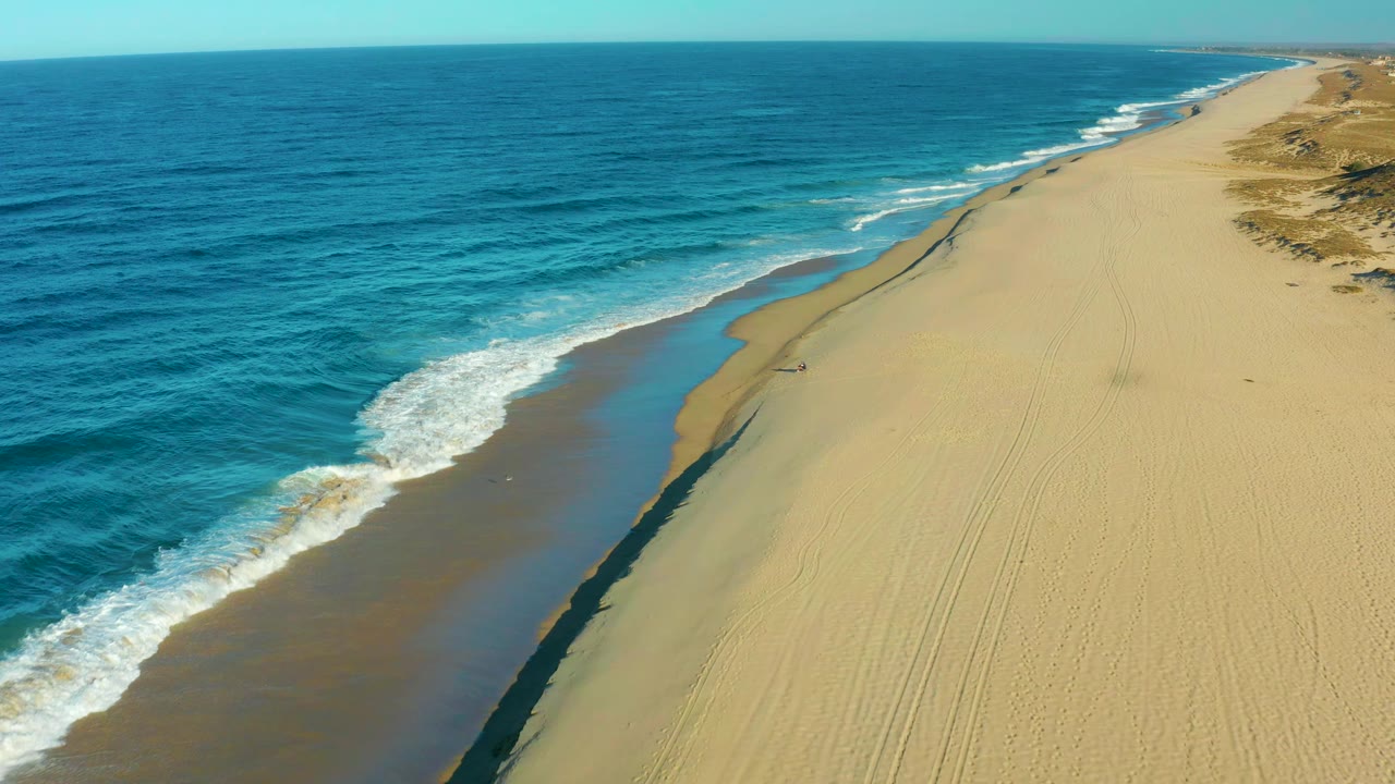 White surf curls toward a broad sandy shore with a tiny person on the beach.