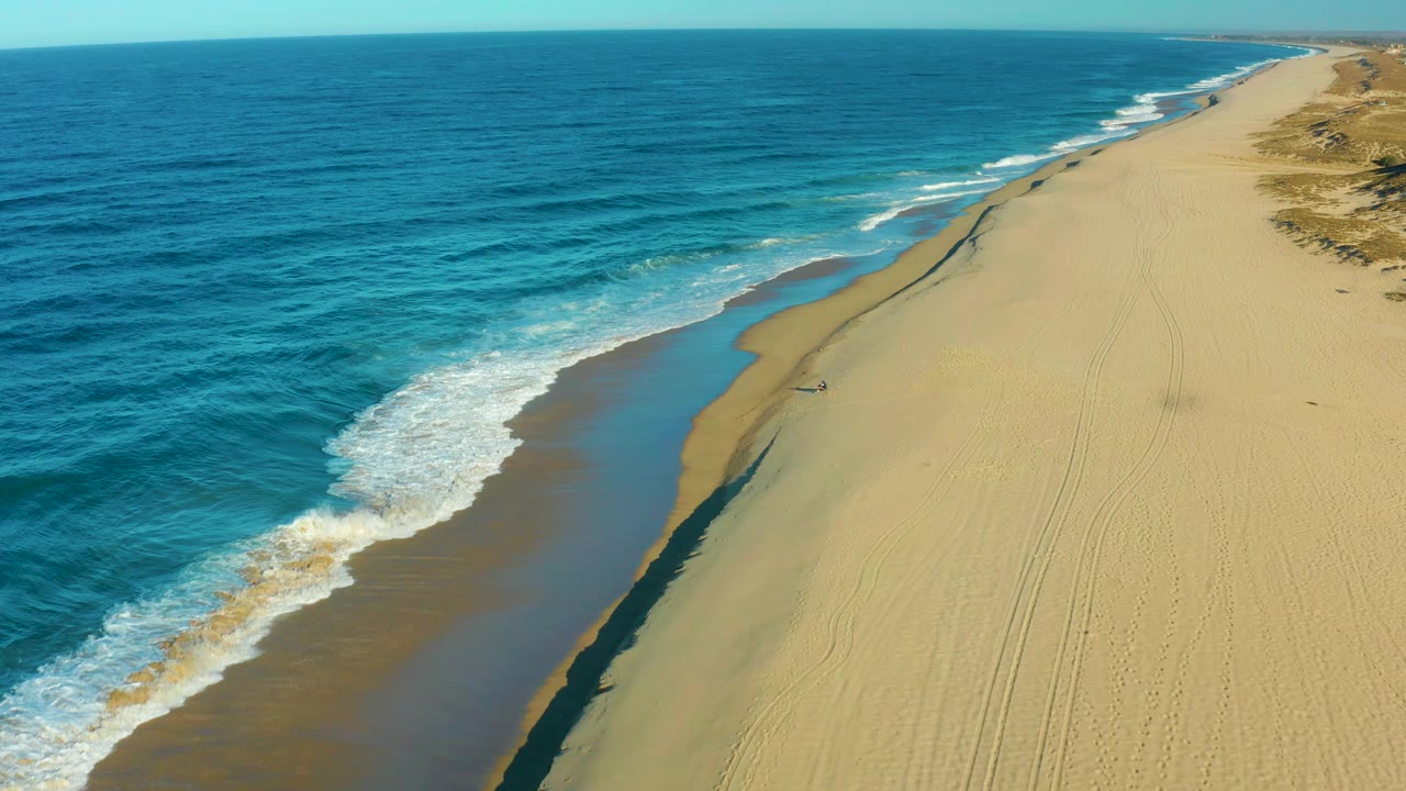 Aerial coastal view of Todos Santos with blue water, a bright white wave line, long tan beach tracks, and one distant person walking near the shore.