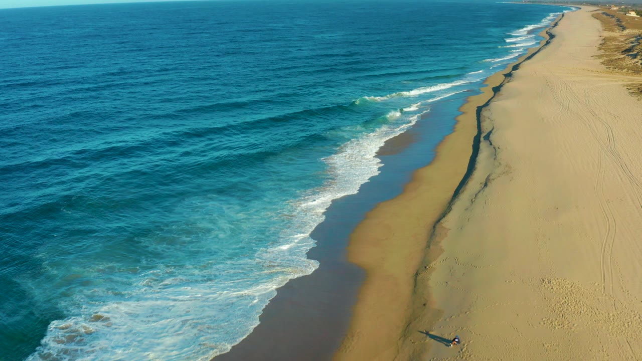 Aerial frame over Todos Santos, Baja California Sur, showing blue ocean on the left, white surf curling onto beige sand, a dark wet line along the beach, and no visible people.